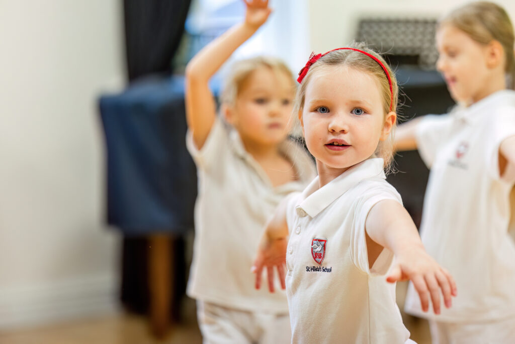 Young girl with a red headband extends her arms forward during a group activity in a hallway; others in white uniforms participate in the background.