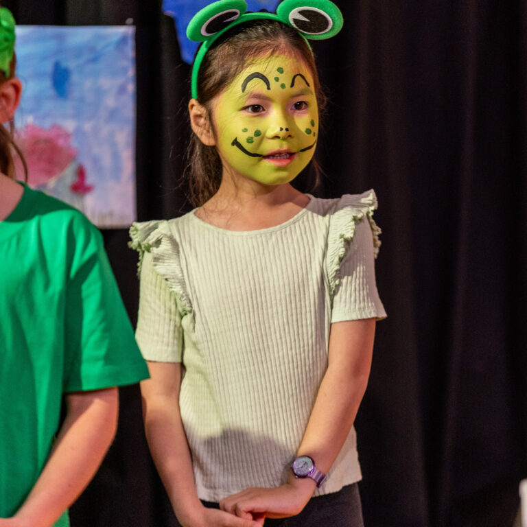 Girl with frog face paint and a green frog-ear headband on stage, hands clasped in front of her.