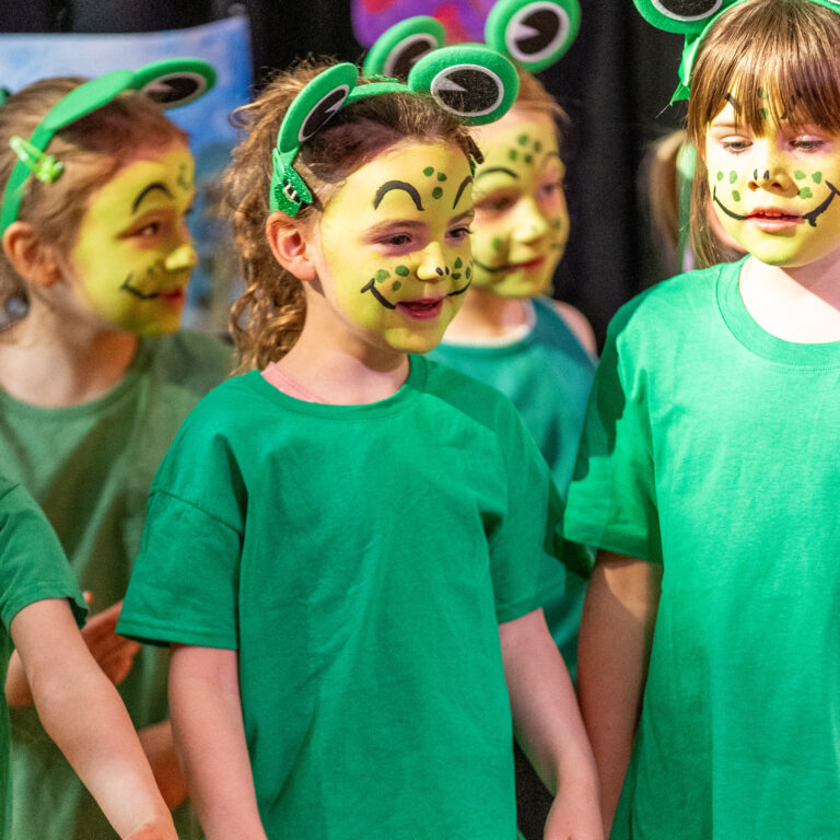 Group of children in green shirts with frog headbands and face paint on stage, smiling and looking around.