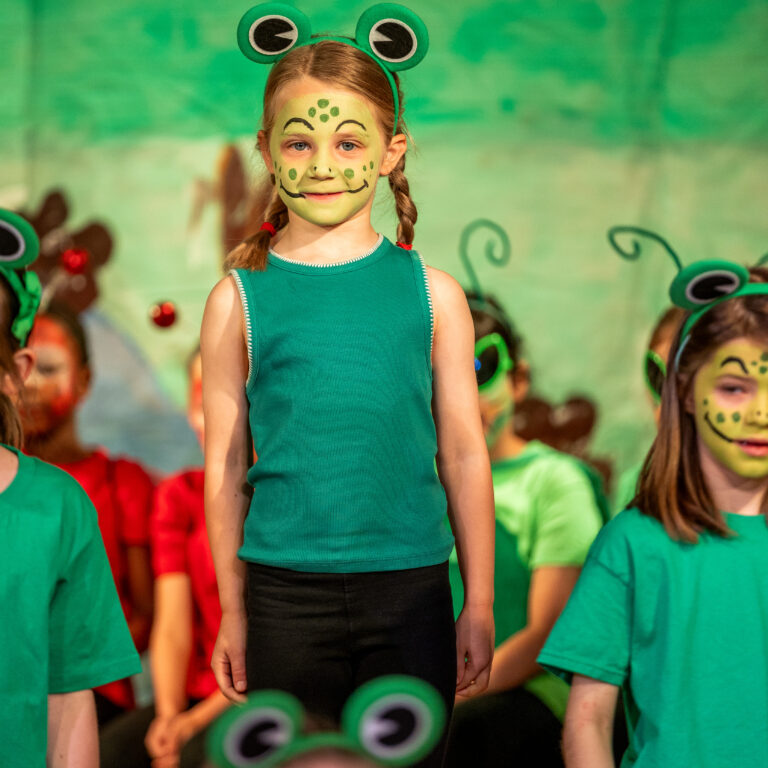 Young girl with frog face paint and a green frog headband stands at the front of a group on a stage during a performance.