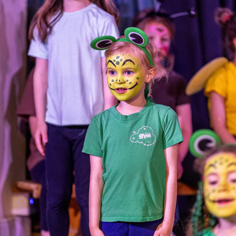 Girl with frog face paint and headband on stage, wearing a green shirt, in a group performance.