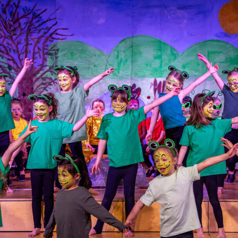 Group of children in frog costumes dancing on a stage, green shirts and headbands with frog eyes, colorful backdrop of hills and sun behind them.