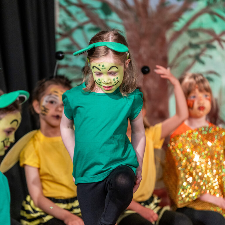 Girl in a green shirt and matching headband steps forward on a stage, her face painted green with dots; background shows other kids in colorful costumes and a forest backdrop.