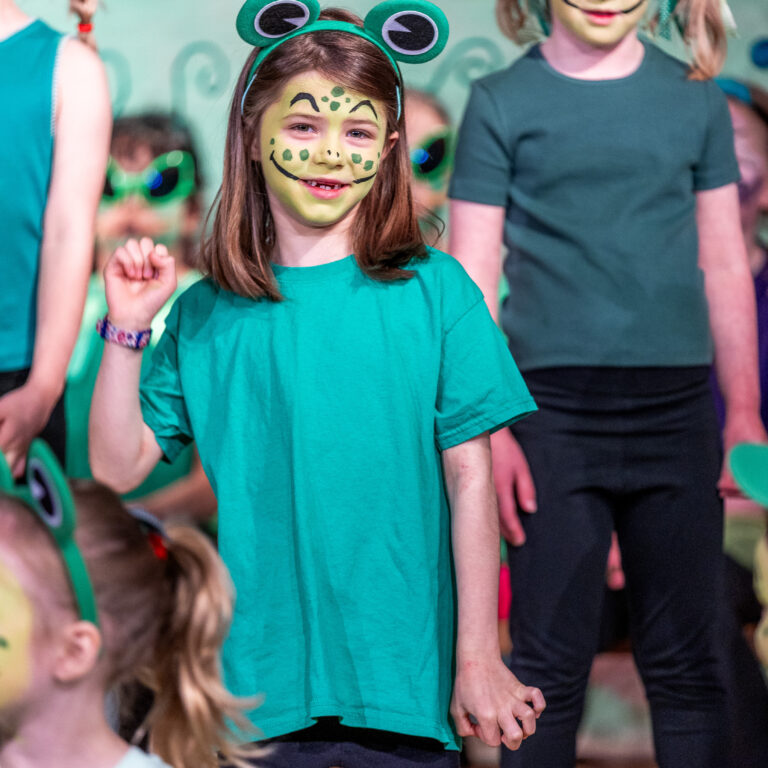 Young girl with frog face paint and a green frog headband smiling at a group of kids in green outfits.