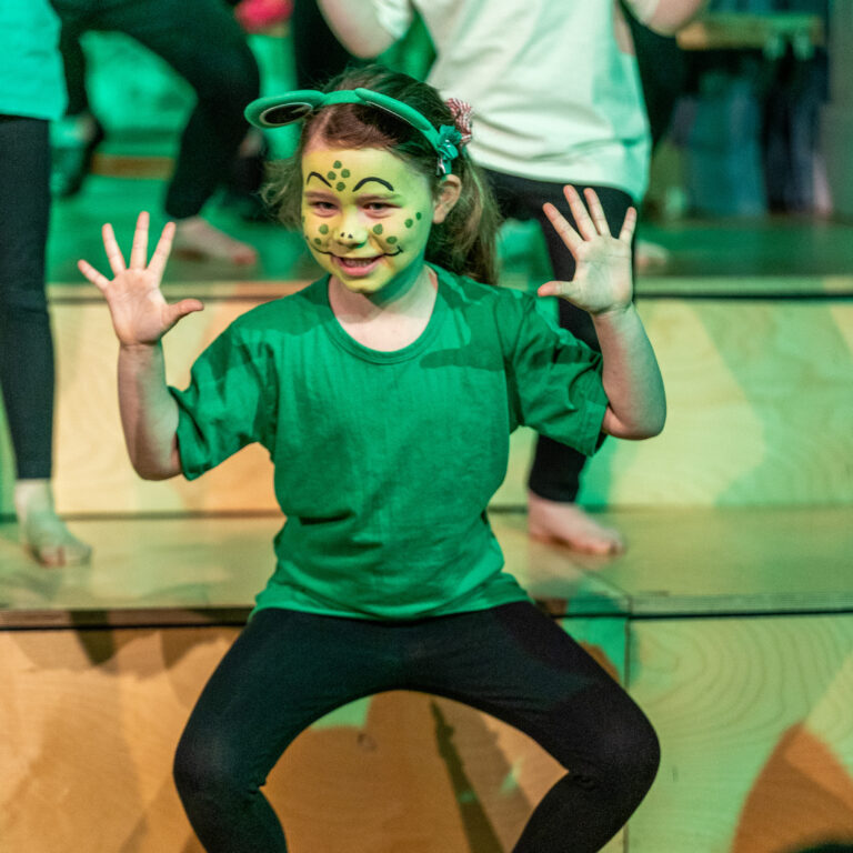 Face-painted girl in a green shirt smiles with hands up, dancing on a wooden stage while other kids lean in the background.