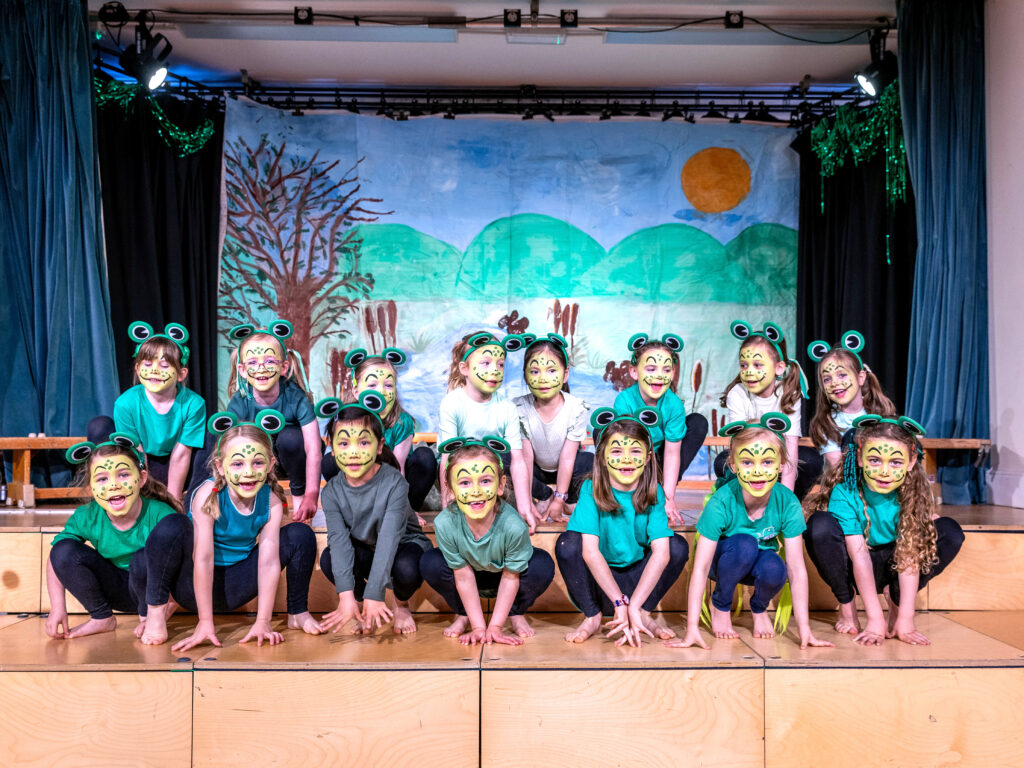 Group of children wearing frog masks and green shirts posing on a stage with a nature-scene backdrop and blue curtains on either side, stage lights above.