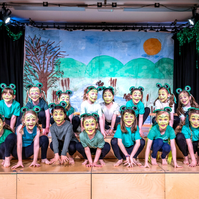 Group of children wearing frog masks and green shirts posing on a stage with a nature-scene backdrop and blue curtains on either side, stage lights above.