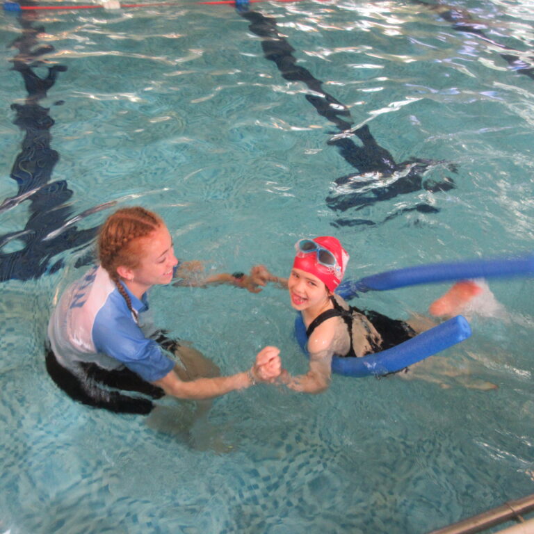 Children in a pool learning to swim, smiling as they practice with a blue kickboard nearby.