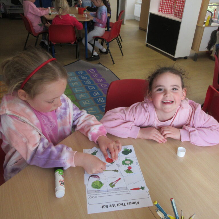 Children sit at a classroom table doing a leaf/vegetable craft, with cards and glue on the table.