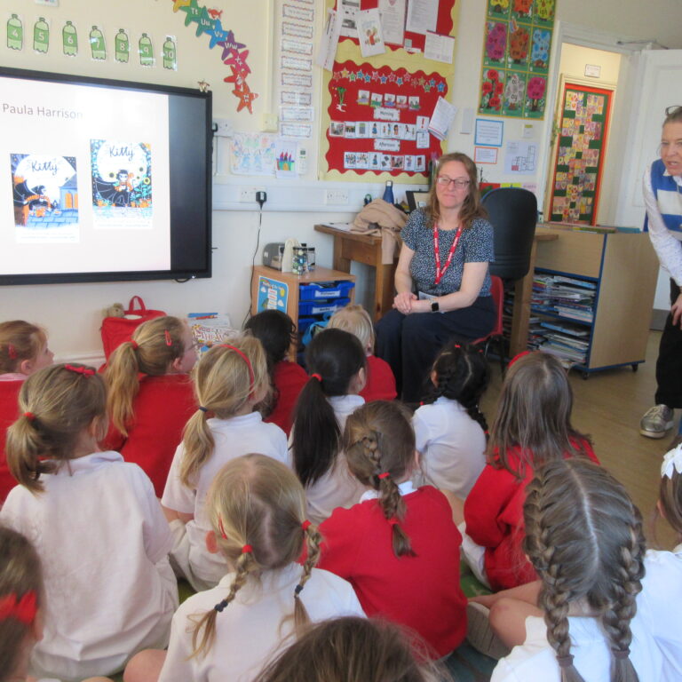 Group of young students in red and white uniforms seated on the floor, listening to two teachers in a classroom with a large presentation screen showing 'Paula Harrison' and book covers.