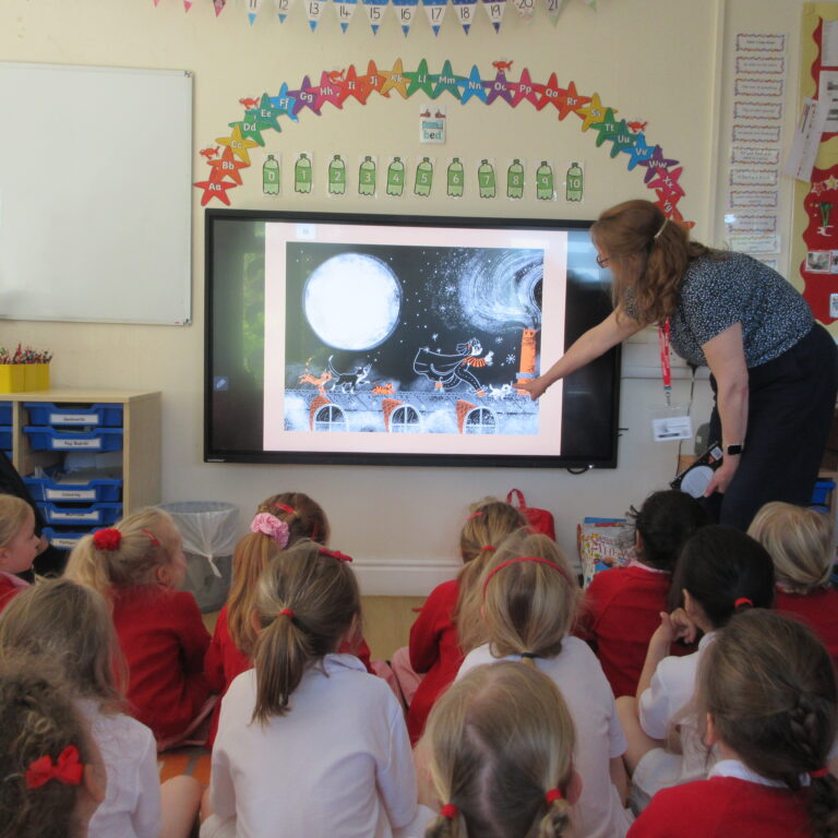 Teacher points to a picture on a large interactive screen as young students sit on the floor watching a lesson in a classroom lighted room.