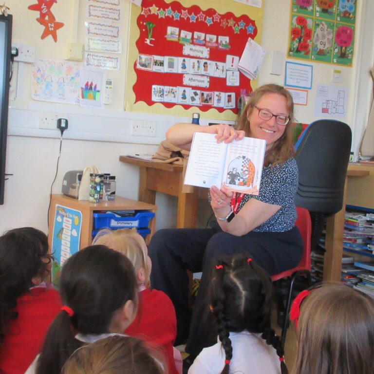 Teacher reading a storybook to a group of children in a classroom, colorful student posters on the wall behind.