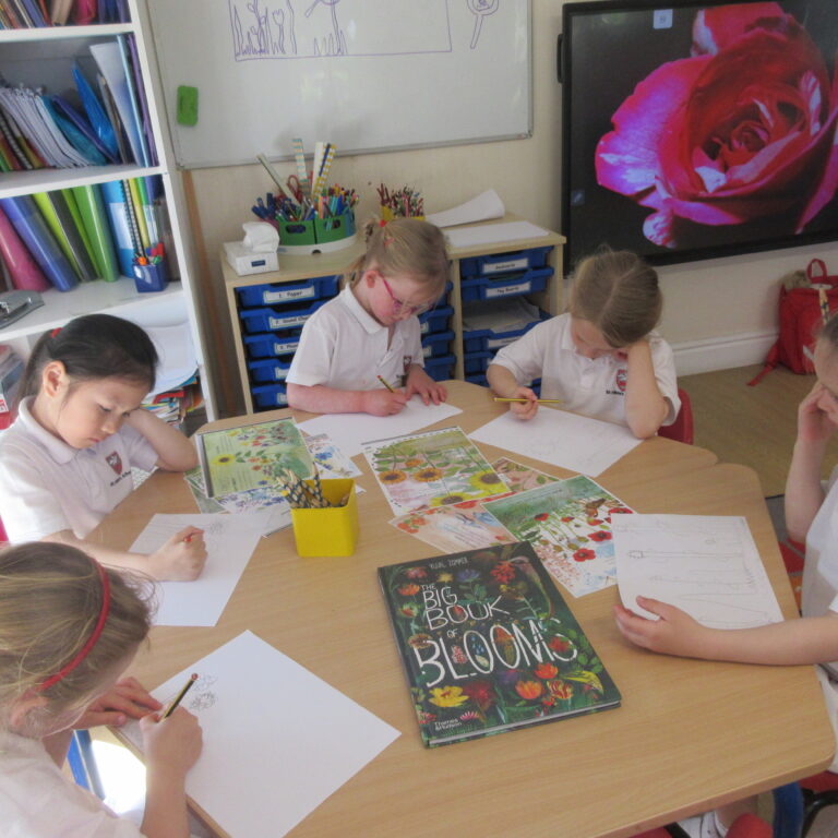 Group of children in white shirts drawing at a classroom table with art supplies nearby and colorful folders on shelves in the background.