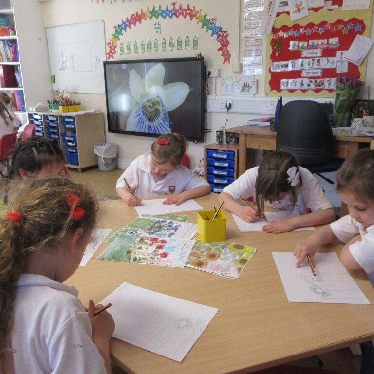 Children in white uniforms sit around a tables drawing with pencils in a colorful classroom show of plants and posters in the background.