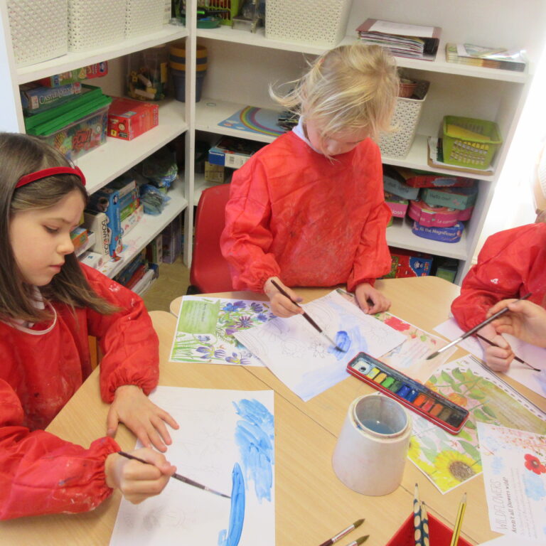 Group of children in red smocks working on colorful paintings at a classroom table with shelves of activities in the background.