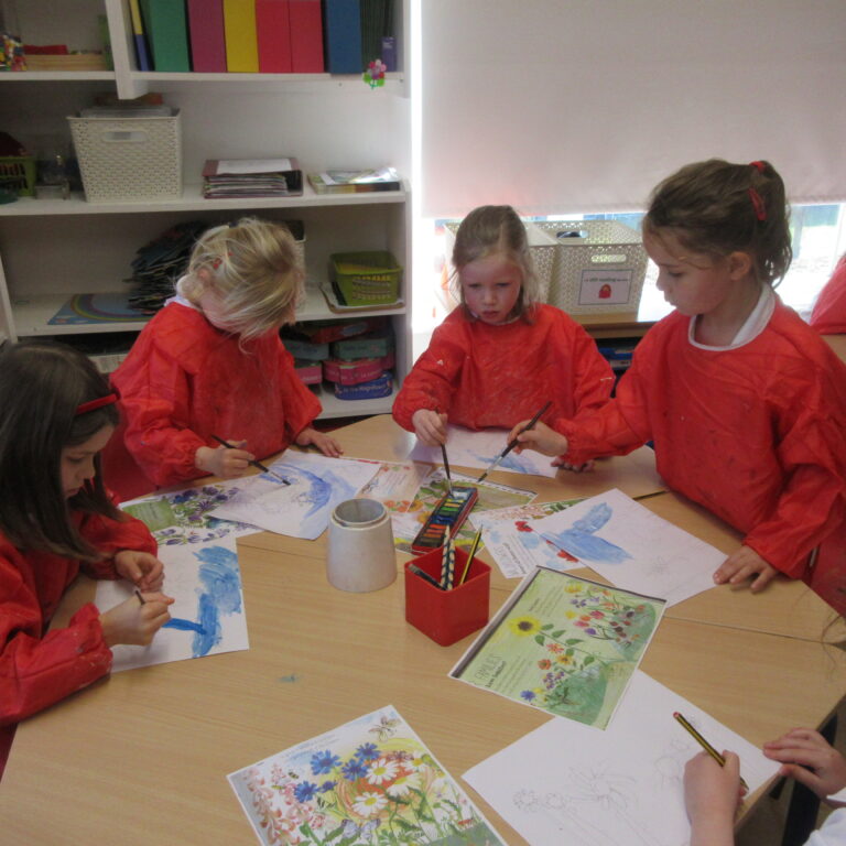 Group of young children in red smocks painting at a classroom table, with art supplies and colorful worksheets nearby.