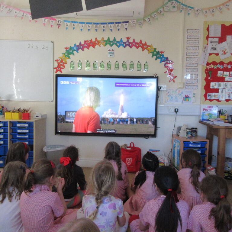 Group of young students seated on the floor in pink uniforms, facing a large classroom TV screen showing a video.