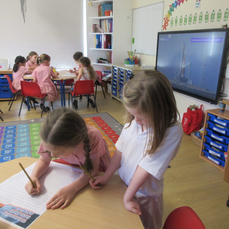 Several young students sit at round tables, writing in notebooks during a classroom activity with colorful floor mat and educational posters in the background.