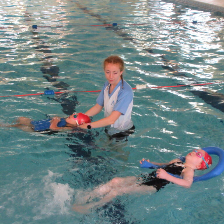 Instructor in blue shirt helping a child with a red swim cap float in a pool lined with lane dividers.