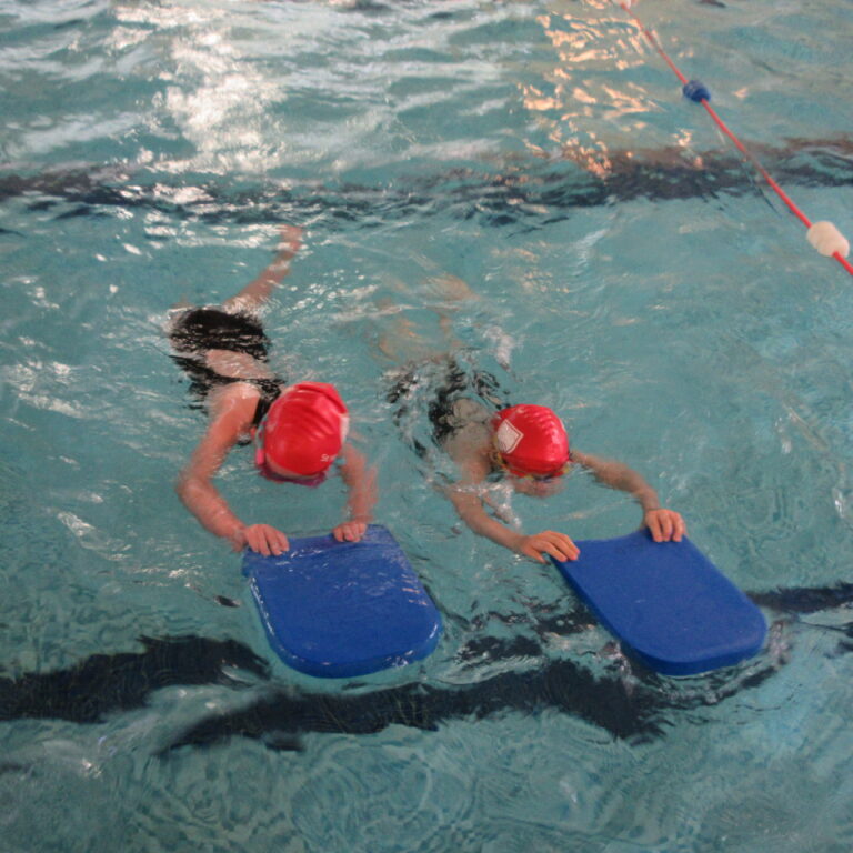Two swimmers wearing red swim caps use blue kickboards in a pool during a training session
