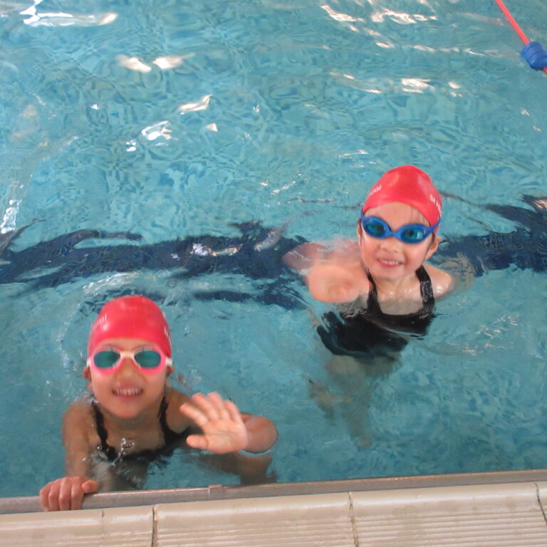Two children in a swimming pool wearing red swim caps and goggles, smiling at the camera by the pool edge.