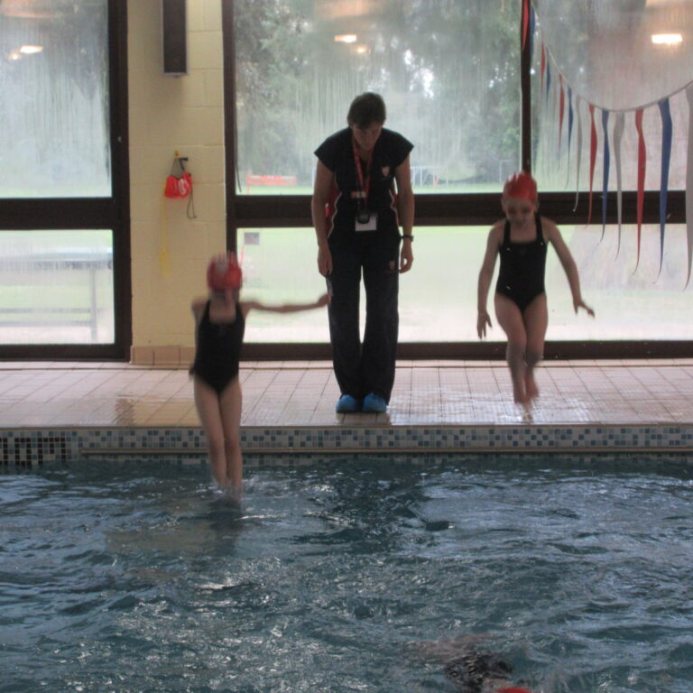 Child in a black swimsuit jumps into a pool as another swimmer and a coach stand at the edge indoors, with decorative banners in the background.