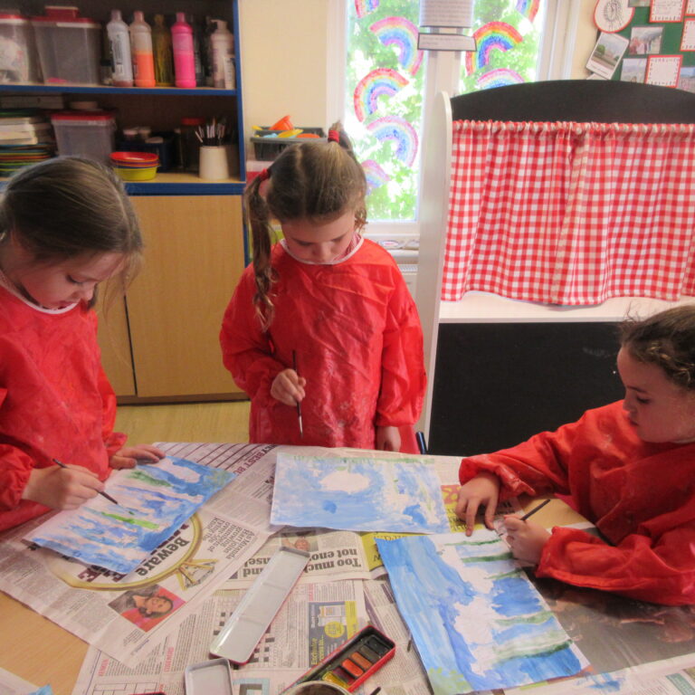 Children in red smocks painting blue watercolors at a table covered with newspapers in a bright classroom.