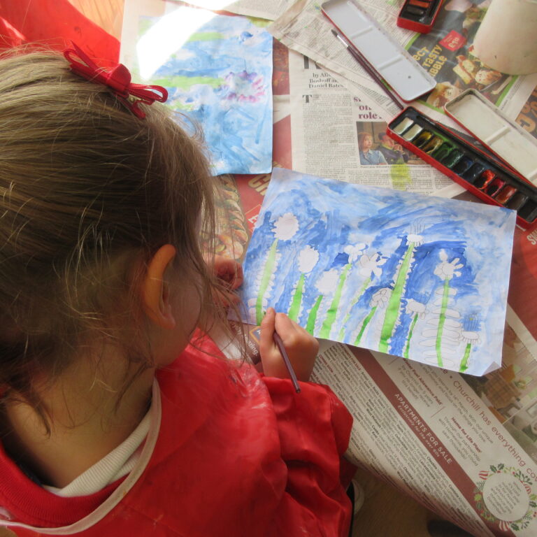 Child in a red apron paints blue flowers with watercolor on a sheet of paper laid over newspapers.