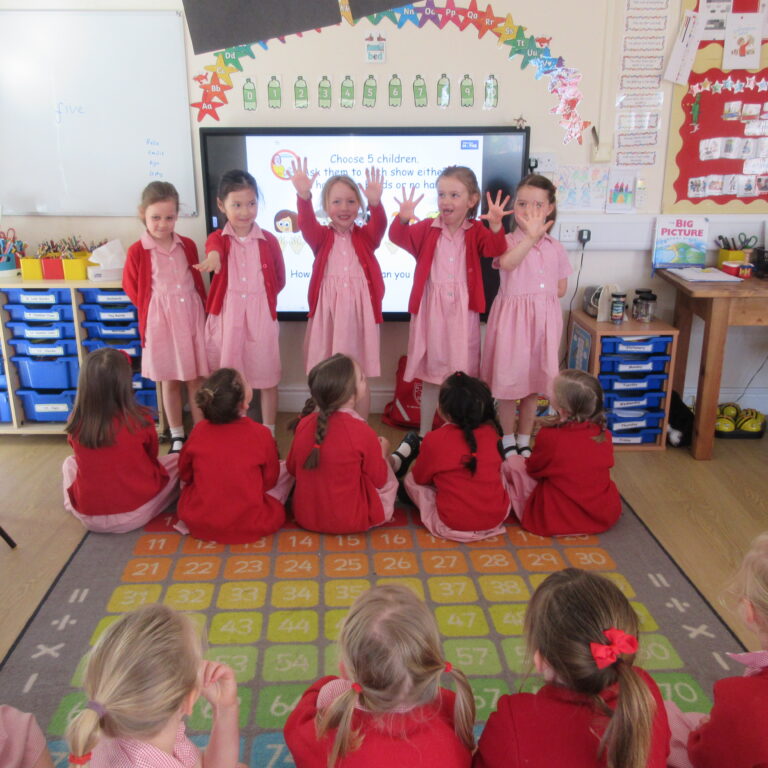 Five girls in red uniforms stand at the front of a classroom with a digital display, hands raised, while classmates sit on a colorful number rug.