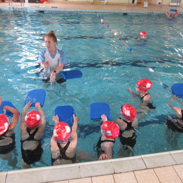 Swimming class in an indoor pool: instructor guiding children with blue kickboards during a lesson, all wearing red swim caps.