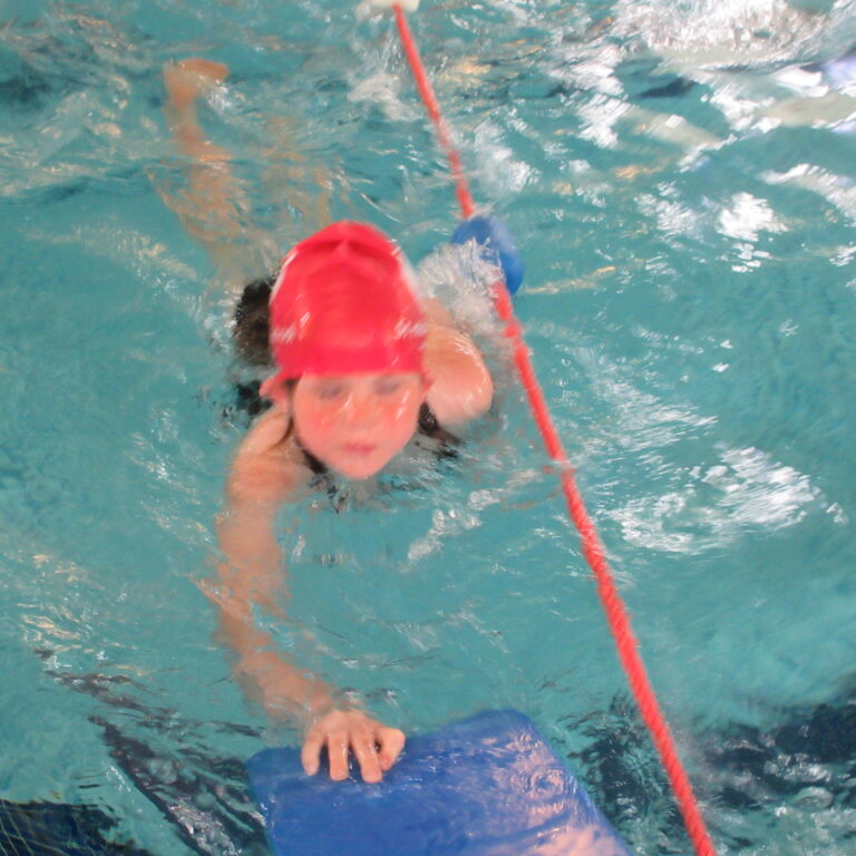 Child with pink swim cap holds a blue kickboard in a pool during a swim lesson, with a red lane line nearby/scrim of water.