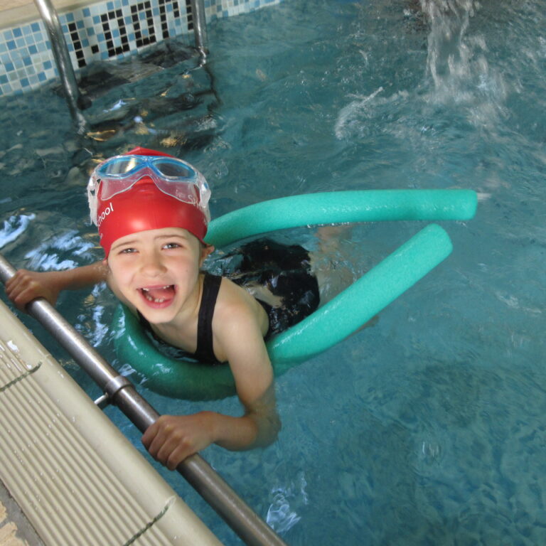 Child wearing a red swim cap and goggles smiles while holding the pool edge, with green pool noodles nearby in a blue-tiled pool area.