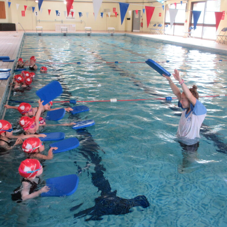 Indoor swimming pool lesson: children with red swim caps using blue kickboards, coach at pool edge giving instructions.