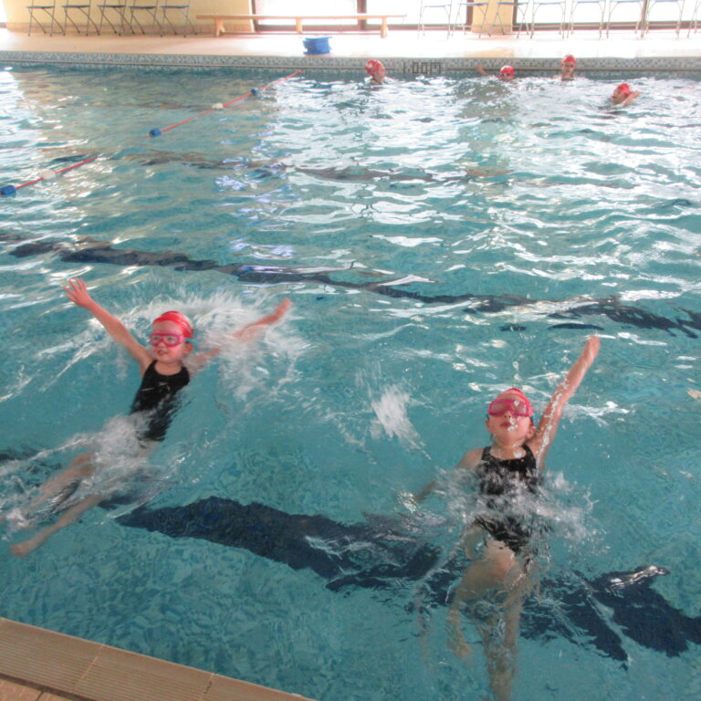 Children wearing red swim caps swimming in an indoor pool during a lesson, with lane lines visible on the water surface