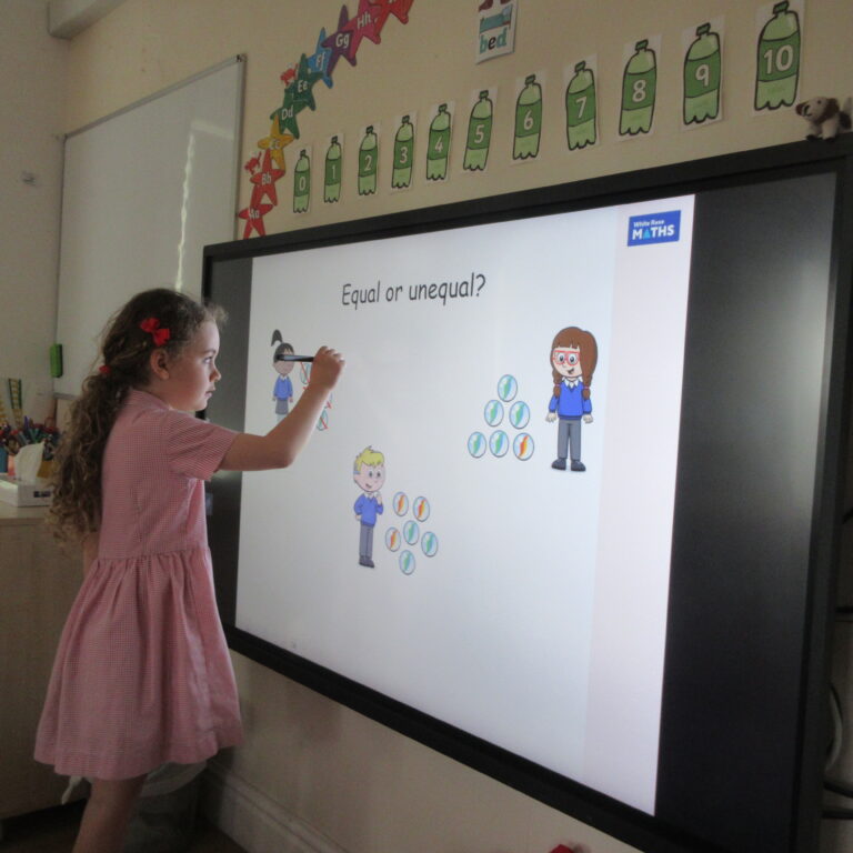 Girl in a pink dress writes on an interactive whiteboard showing a math activity: 'Equal or unequal?' with cartoon characters and shapes.
