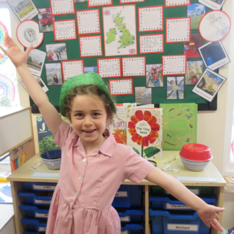 Smiling girl in a pink checkered dress raises one arm in a classroom with a green bulletin board and colorful posters behind her.