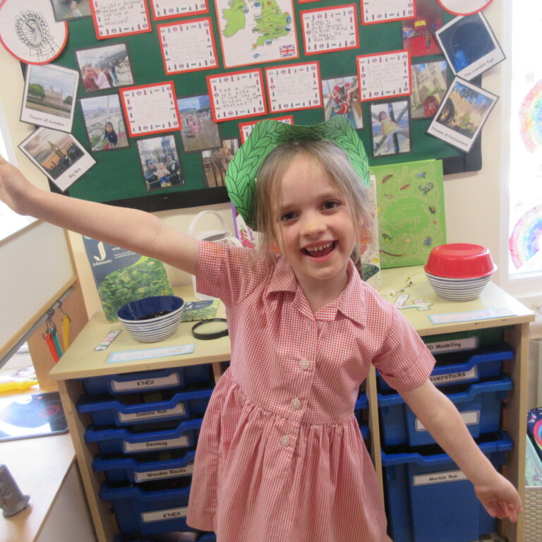 Young girl in a red check dress stands in a classroom with arms outstretched, rainbow drawings on the windows and a bulletin board of photos behind her.