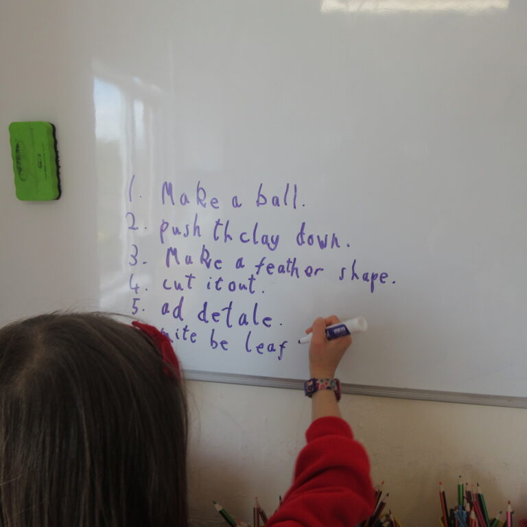 Whiteboard with a handwritten list of classroom tasks; a child in red writes with a marker, green eraser on the left.