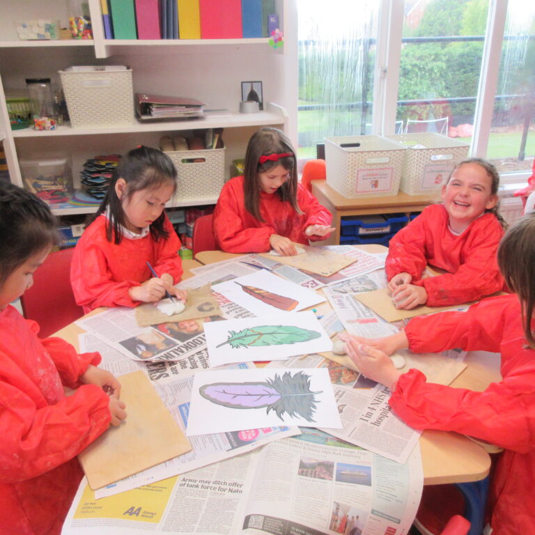 Group of children in red smocks drawing and painting leaf prints on a newspaper-covered table in a bright classroom.