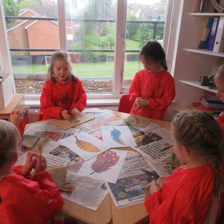 Children in red smocks sit around a table covered in newspapers, crafting leaf-print artworks with paint in a classroom.