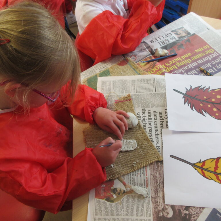 Child with braided hair wearing a red smock, shaping white clay on burlap while newspapers and a feather illustration lie on the table.