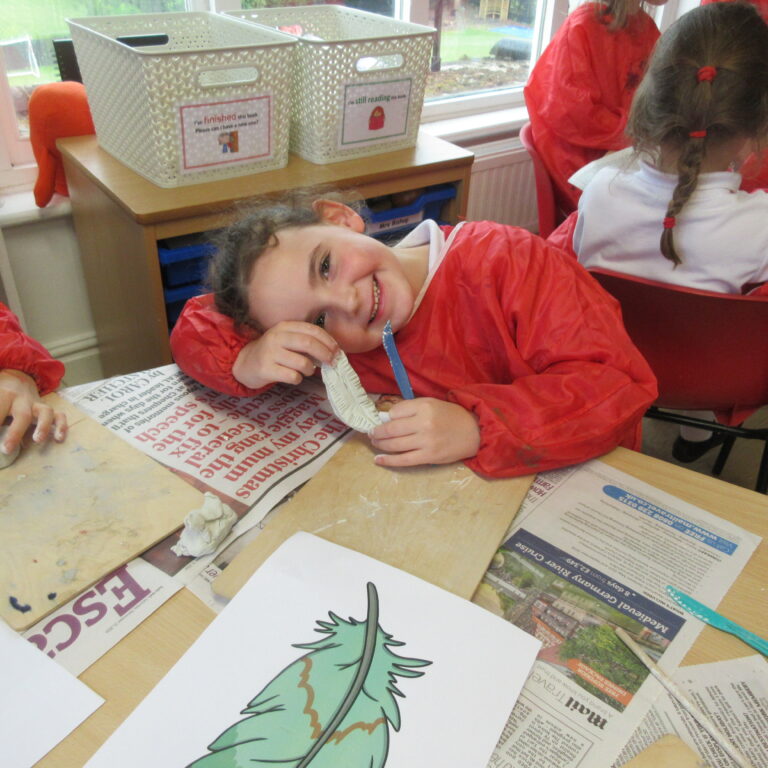 Child in a red smock smiling while shaping clay at a desk covered with newspapers in a classroom area
