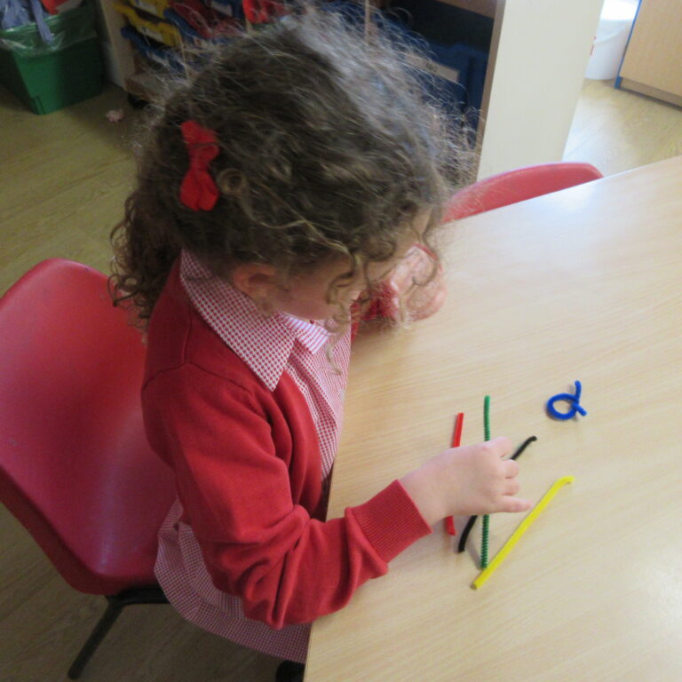 Child in red cardigan sits at a table, shaping colorful pipe cleaners for a craft activity in a classroom.