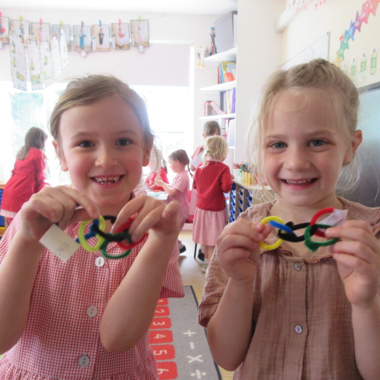 Two smiling young girls in a classroom showing colorful pipe-cleaner bracelets they made together on a craft day.