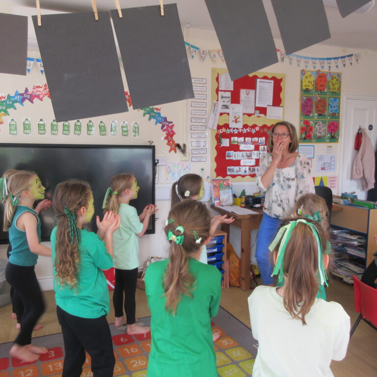 A teacher at the front of a classroom leading a group of children in a clapping activity; students wear green shirts and some have face paint.