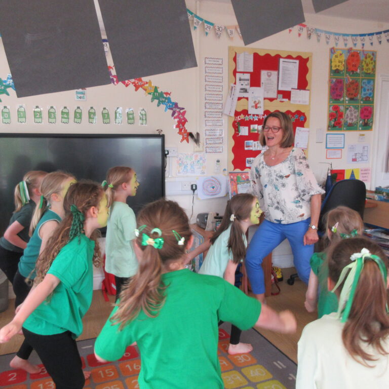 Group of young students in green shirts holding hands and dancing in a classroom as a teacher leads the activity.