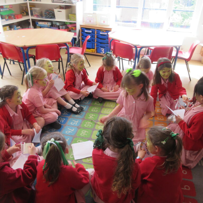 Group of young children in red uniforms seated in a circle on a colorful number rug, with face paint, in a bright classroom.