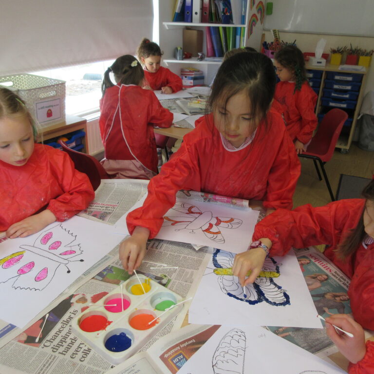 Children in red smocks drawing and painting at a table covered with newspaper in a classroom setting.