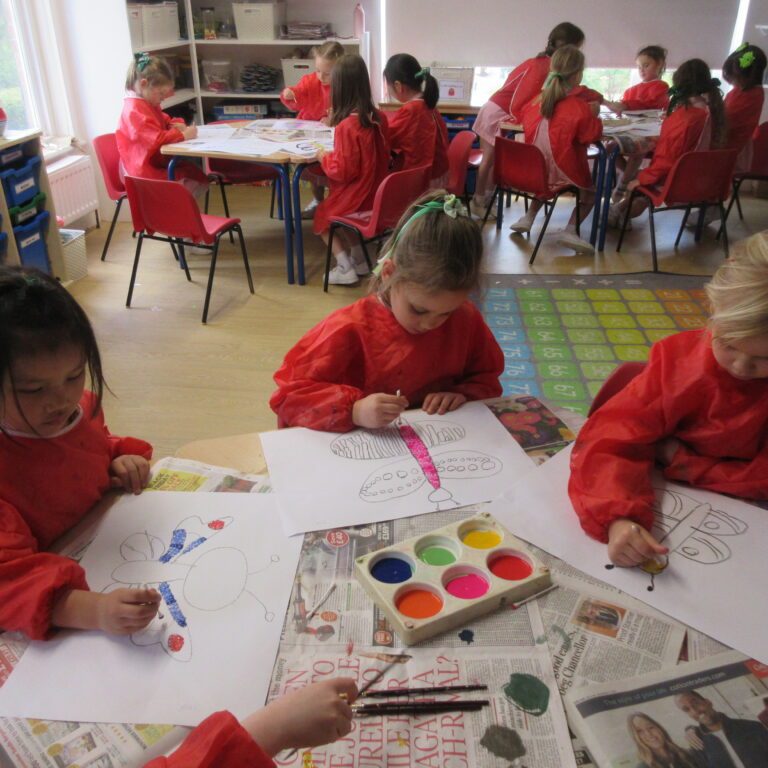 Group of children in red smocks painting at tables in a bright classroom with newspapers on the table