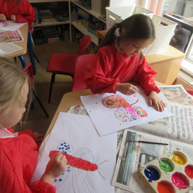 Children in red aprons are painting colorful circular designs on white paper at a classroom table with a paint palette nearby.